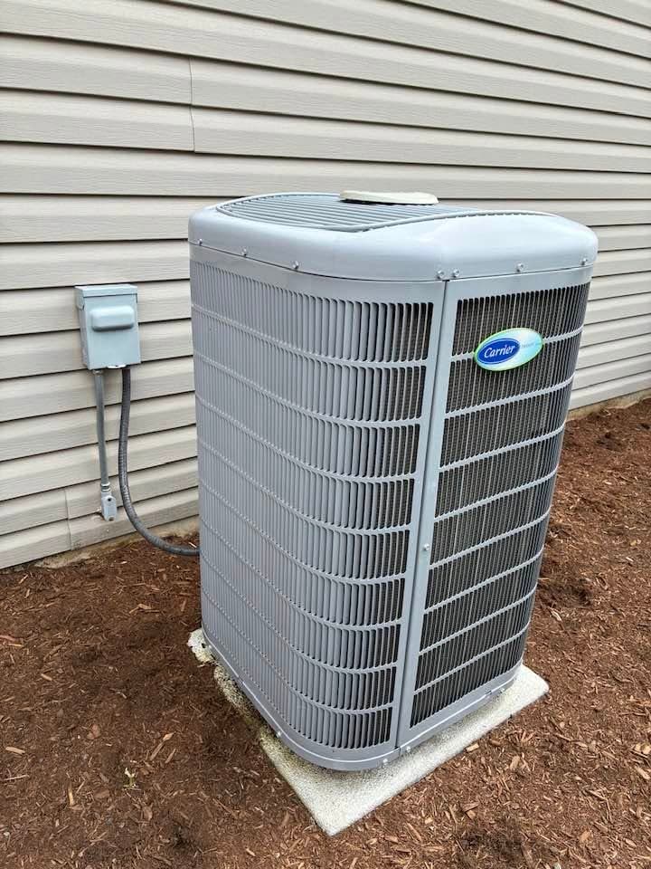 Carrier central air conditioner condenser on a concrete pad beside a house, with the disconnect box and conduit on the siding